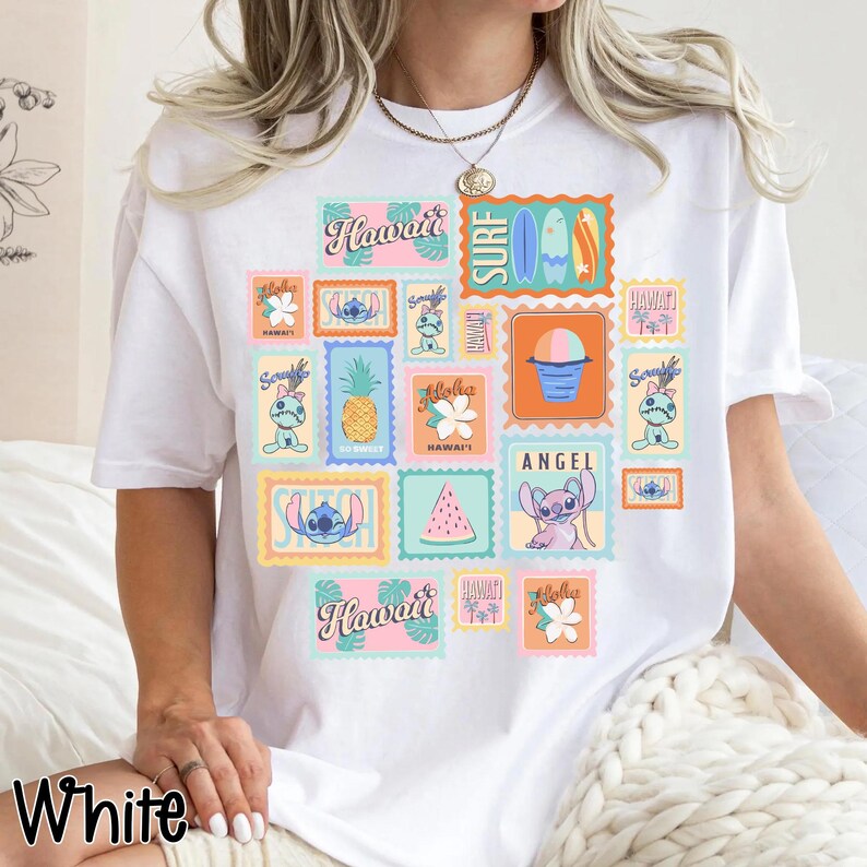 May include: White t-shirt featuring a colorful collage of Hawaiian-themed postage stamp designs. The stamps include images of surfboards, pineapples, flowers, and the character Stitch. The word "Hawaii" is prominently displayed on several stamps.
