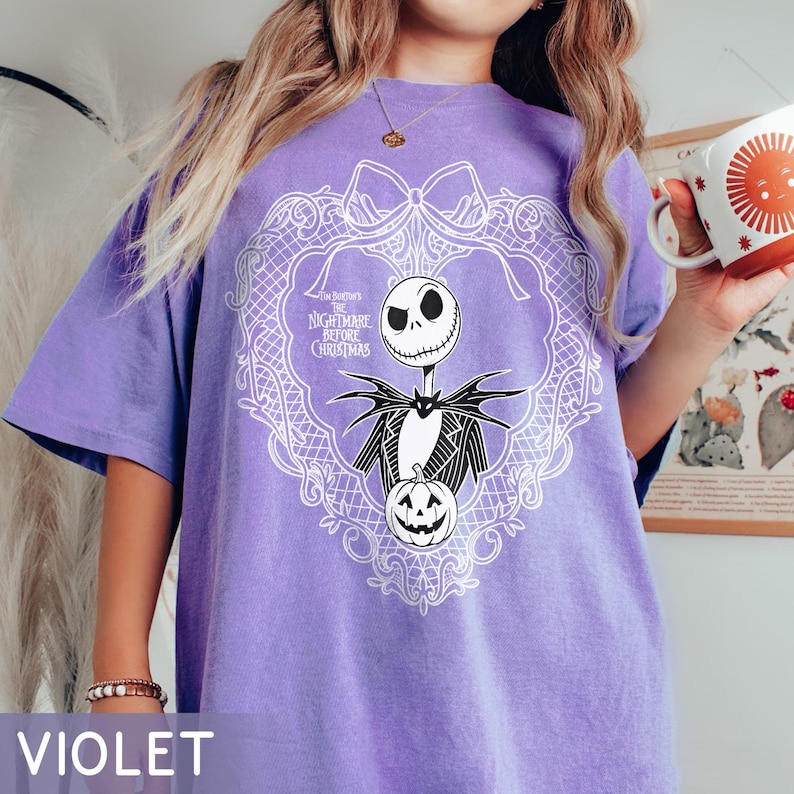 May include: A violet t-shirt with a white graphic of Jack Skellington from "The Nightmare Before Christmas." The design includes a decorative frame, a pumpkin, and the movie title. The shirt is a solid violet colour.