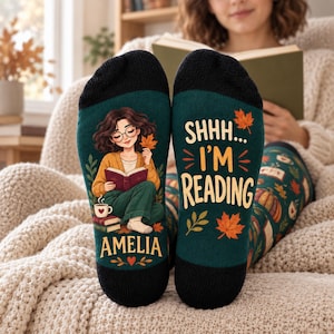 May include: A pair of dark green socks featuring a black cuff and toe. One sock displays an illustration of a person reading with a coffee mug and the name AMELIA. The other sock features the text SHHH... I'M READING with orange autumn leaves.