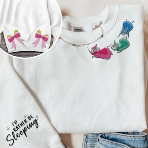May include: White sweatshirt with a black graphic that says "I'd rather be sleeping" on the left sleeve. The right sleeve has two pink bows. The front of the sweatshirt has three fairies flying with wands.
