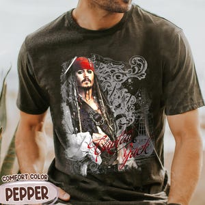May include: Dark grey Comfort Color t-shirt featuring a graphic of a pirate with a red bandana and the text "Captain Jack". The design includes a ship and decorative swirls. The shirt is pepper colored.