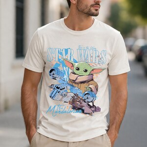 May include: Cream-colored t-shirt featuring a graphic of Grogu from The Mandalorian, with the text "This is the Way" and "Star Wars" in blue. The design includes a blue spaceship and other elements.