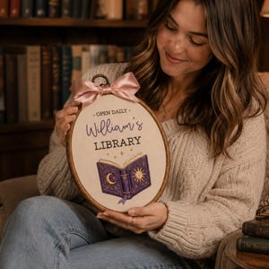 May include: An oval embroidery hoop featuring a cream-colored fabric with the text "OPEN DAILY" and "William's LIBRARY" in purple and black. Below the text is a purple book illustration with a gold moon and sun, topped with a pink ribbon.
