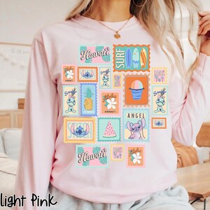 May include: Light pink long-sleeve sweatshirt featuring a colorful collage of Hawaiian-themed postage stamps. The stamps include images of Stitch, pineapples, surfboards, and the word "Hawaii". The text "Light Pink" is at the bottom.