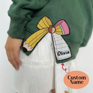 May include: Green sweatshirt with a yellow, pink, and white pencil and bow applique. The applique has a red apple and the text "Olivia" on a white paper.