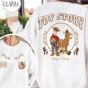 May include: White sweatshirt featuring a "Toy Story" graphic. The design includes Woody and Bullseye, surrounded by a rope circle and cacti. The sweatshirt also has a small design with the number "95" and a horseshoe.