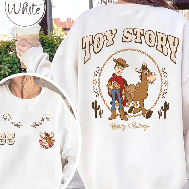 May include: White sweatshirt featuring a "Toy Story" graphic. The design includes Woody and Bullseye, surrounded by a rope circle and cacti. The sweatshirt also has a small design with the number "95" and a horseshoe.