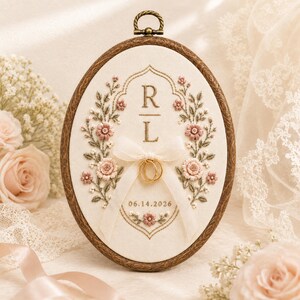 May include: An oval embroidery hoop frame containing cream fabric with floral stitching in pink and green. The center features the letters R and L, a white ribbon holding two gold rings, and the date 06.14.2026 embroidered in brown thread.