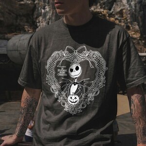 May include: Dark grey t-shirt with a white graphic of Jack Skellington from "The Nightmare Before Christmas." The design includes a decorative frame, a bow, and a pumpkin. The text "The Nightmare Before Christmas" is also visible.