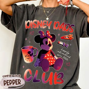 Może przedstawiać: Ciemnoszara koszulka Comfort Color z grafiką Myszki Minnie. Projekt zawiera tekst "Disney Dads Club" i "happily outnumbered since 2026". Na koszulce znajduje się r&oacute;wnież kubek do kawy i spinki do włos&oacute;w.