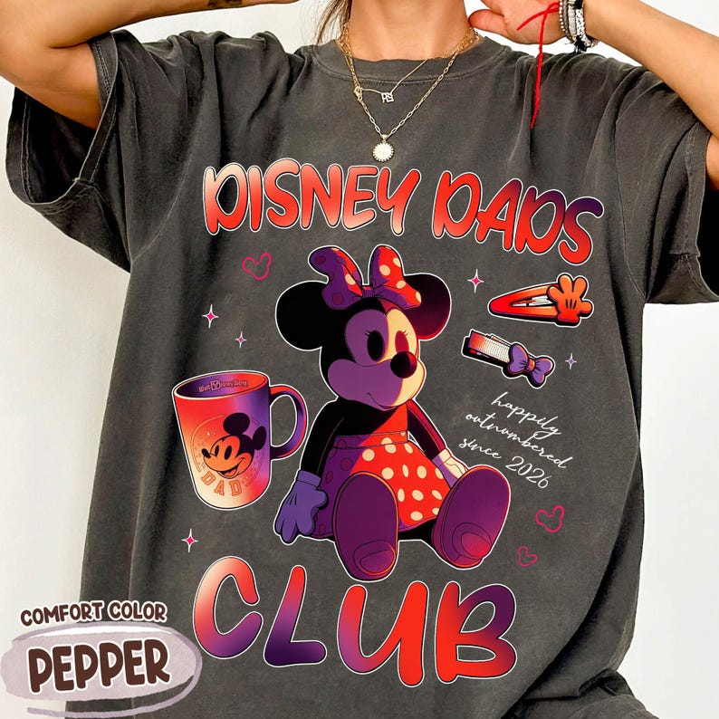 Może przedstawiać: Ciemnoszara koszulka Comfort Color z grafiką Myszki Minnie. Projekt zawiera tekst "Disney Dads Club" i "happily outnumbered since 2026". Na koszulce znajduje się r&oacute;wnież kubek do kawy i spinki do włos&oacute;w.