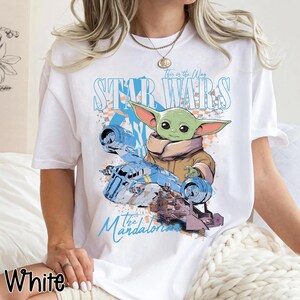 May include: White t-shirt featuring a graphic of Grogu from The Mandalorian and a blue ship. The text "This is the Way" and "Star Wars" are above the image, and "The Mandalorian" is below. The word "White" is at the bottom left.