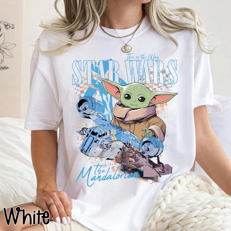 May include: White t-shirt featuring a graphic of Grogu from The Mandalorian and a blue ship. The text "This is the Way" and "Star Wars" are above the image, and "The Mandalorian" is below. The word "White" is at the bottom left.