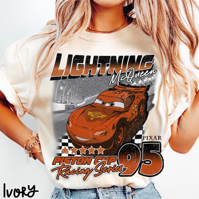 May include: Ivory-colored t-shirt featuring a graphic of Lightning McQueen from the Pixar movie Cars. The design includes the text "LIGHTNING McQueen", "PISTON CUP Racing Series", and the number "95". The shirt has a vintage, distressed look.