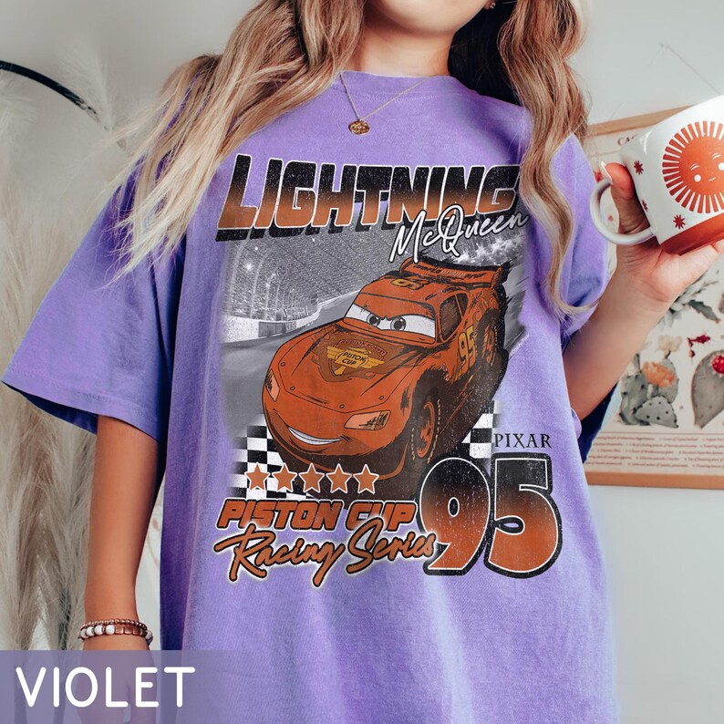 May include: Violet t-shirt featuring a graphic of Lightning McQueen from the movie Cars. The design includes the text "LIGHTNING McQueen", "PISTON CUP Racing Series", and the number "95". The shirt has a vintage, distressed look.