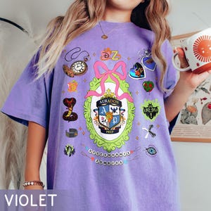 Może przedstawiać: Fioletowa koszulka z kolorowym nadrukiem graficznym przedstawiającym herb, kokardę i inne motywy tematyczne. Koszulka ma luźny kr&oacute;j i jest wykonana z miękkiego materiału.