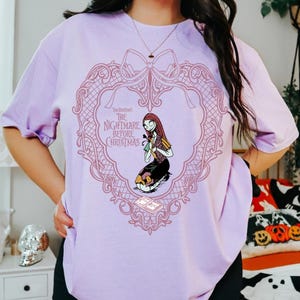 May include: A lilac t-shirt with a pink heart-shaped frame design. The text reads "Tim Burton's The Nightmare Before Christmas." Inside the frame is a cartoon illustration of Sally. The shirt has short sleeves.