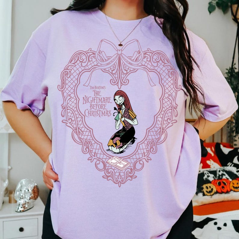 May include: A lilac t-shirt with a pink heart-shaped frame design. The text reads "Tim Burton's The Nightmare Before Christmas." Inside the frame is a cartoon illustration of Sally. The shirt has short sleeves.