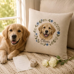 May include: A square beige throw pillow featuring an embroidered portrait of a golden retriever dog centered within a floral wreath of blue flowers and green leaves, with the name Oliver stitched in cursive script below the dog's portrait.