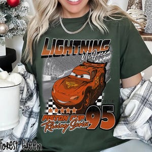 May include: Forest green t-shirt featuring Lightning McQueen from the movie Cars. The design includes the text "Lightning McQueen", "Piston Cup Racing Series", and the number "95". The graphic is in orange, white, and black.