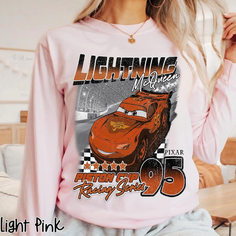 May include: Light pink long-sleeve sweatshirt featuring a graphic of Lightning McQueen from the Pixar Cars movie. The design includes the text "LIGHTNING McQueen", "PISTON CUP Racing Series", and the number "95".