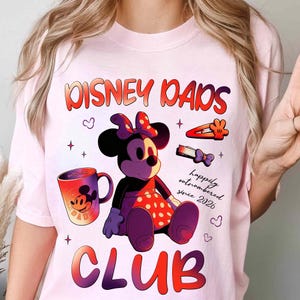 Może przedstawiać: Jasnor&oacute;żowa koszulka z grafiką przedstawiającą Minnie Mouse, kubek do kawy i napisem "Disney Dads Club". Projekt zawiera frazę "happily outnumbered since 2026."