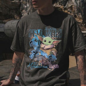 May include: Dark gray t-shirt featuring a graphic of Grogu from The Mandalorian, with the text "Star Wars" and "This is the Way". The design includes a blue spaceship and Grogu in a tan coat.