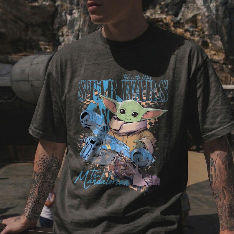 May include: Dark gray t-shirt featuring a graphic of Grogu from The Mandalorian, with the text "Star Wars" and "This is the Way". The design includes a blue spaceship and Grogu in a tan coat.
