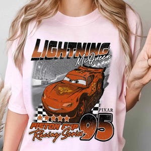 May include: Light pink t-shirt featuring a graphic of Lightning McQueen from the Pixar movie Cars. The design includes the text "LIGHTNING McQueen", "PIXAR", "95", and "Piston Cup Racing Series" in orange and black.