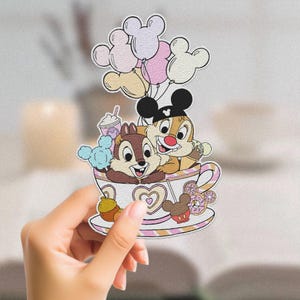 May include: Chip 'n Dale are riding in a pink and white teacup with balloons. The teacup has a heart design and a donut on the side. Chip is wearing a brown and white outfit and Dale is wearing a red and white outfit.