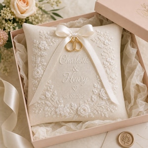 May include: A square cream-colored ring pillow featuring white floral embroidery, a satin ribbon bow, and two gold rings. The text reads Charlotte & Henry with the date 08.09.2026. The pillow is presented in a light pink gift box with tissue paper.