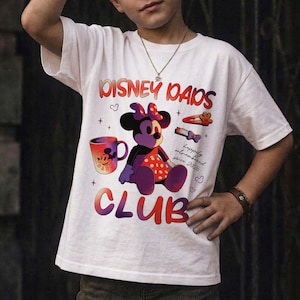 Może przedstawiać: Biała koszulka z grafiką przedstawiającą Minnie Mouse, kubek do kawy i napisem "Disney Dads Club". Projekt zawiera serca i frazę "happily outnumbered since 2020."