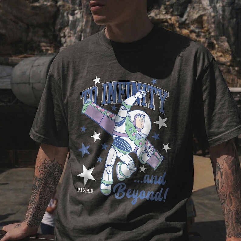 Puede incluir: Camiseta gris oscuro con Buzz Lightyear de Toy Story. El dise&ntilde;o incluye la frase "To Infinity...and Beyond!" en azul y blanco, con estrellas. El logotipo de Pixar est&aacute; en la parte inferior.