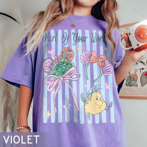 Puede incluir: Una camiseta violeta con un estampado gr&aacute;fico de rayas verticales azul claro y el texto Part Of Your World. El dise&ntilde;o incluye un dulce verde con forma de concha con un lazo rosa, un par de orejas con conchas y un personaje de pez amarillo.