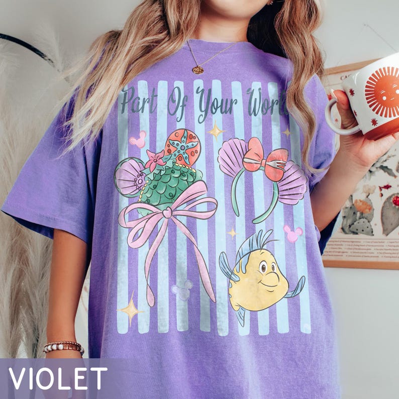 Puede incluir: Una camiseta violeta con un estampado gr&aacute;fico de rayas verticales azul claro y el texto Part Of Your World. El dise&ntilde;o incluye un dulce verde con forma de concha con un lazo rosa, un par de orejas con conchas y un personaje de pez amarillo.