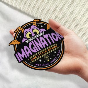 May include: A black and orange circular patch with a purple dragon character. The text on the patch reads "Journey into Imagination, One Little Spark Inspiring Since 1983".