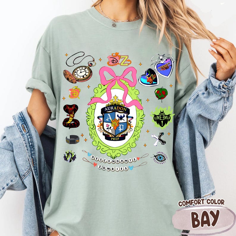 Może przedstawiać: Szałwiowa zielona koszulka z r&oacute;żnymi kolorowymi grafikami, w tym r&oacute;żową kokardą, zegarkiem kieszonkowym i napisem "Descendants Zombies". Koszulka zawiera r&oacute;wnież napis "Auradon Prep".