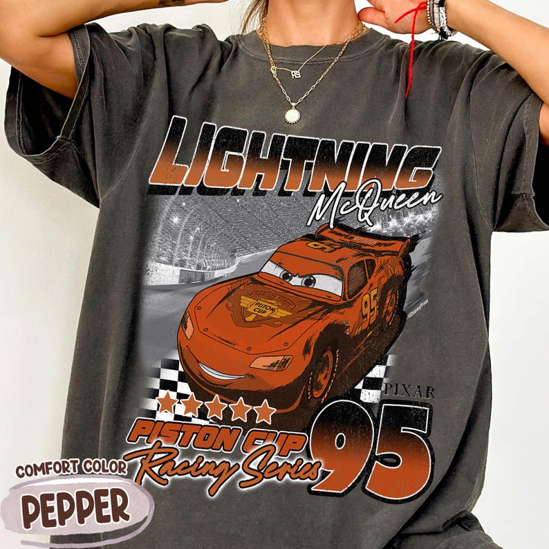 May include: Dark grey t-shirt featuring Lightning McQueen from the movie Cars. The design includes the text "LIGHTNING McQueen", "PISTON CUP Racing Series", and the number "95". The shirt is labeled "Comfort Color Pepper".