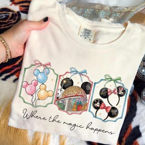 May include: Cream-colored sweatshirt with a colorful graphic featuring Mickey Mouse balloons, a park scene, and Minnie Mouse ears. The text "Where the magic happens" is written in script below the graphic.
