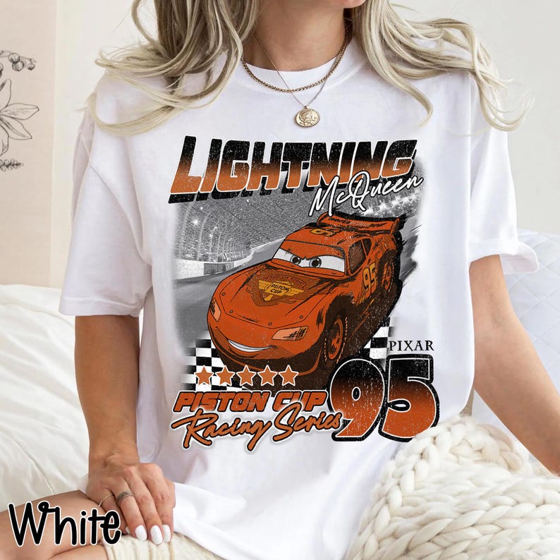 May include: White t-shirt featuring a graphic of Lightning McQueen from the Pixar movie Cars. The design includes the text "Lightning McQueen", "Piston Cup Racing Series", and the number "95". The shirt also has a checkered flag design.