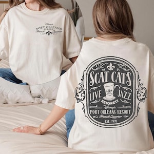 May include: A cream-colored t-shirt featuring a small logo on the front chest that reads Disney's Port Orleans Resort French Quarter. The back displays a large oval graphic with the text Scat Cats Live Jazz Beignets Port Orleans Resort Est. 1991.
