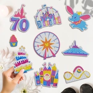May include: A collection of colorful embroidered patches featuring various designs. Included are a castle with "Disneyland" text, a Ferris wheel with a Mickey Mouse icon, a roller coaster, and a flying elephant. Other patches include a cupcake and a birthday cake.