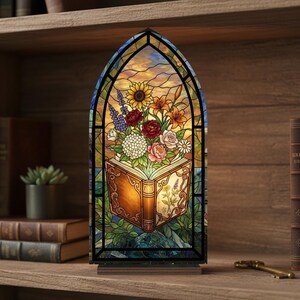 May include: Stained glass lamp in an arched window shape, featuring a colorful floral arrangement emerging from an open book. The flowers include sunflowers, roses, and lilies. The lamp sits on a wooden base.