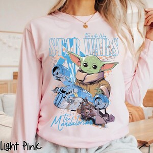 May include: Light pink long-sleeve sweatshirt featuring a graphic of Grogu from The Mandalorian and the text "Star Wars" and "This is the Way". The design includes a light blue spaceship and a brown and tan character.