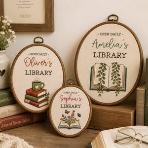 May include: Three oval embroidery hoops featuring personalized library designs on cream fabric. The hoops display the text 'OPEN DAILY' and names 'Oliver's', 'Amelia's', and 'Sophia's' above 'LIBRARY', accompanied by embroidered books, plants, and flowers.