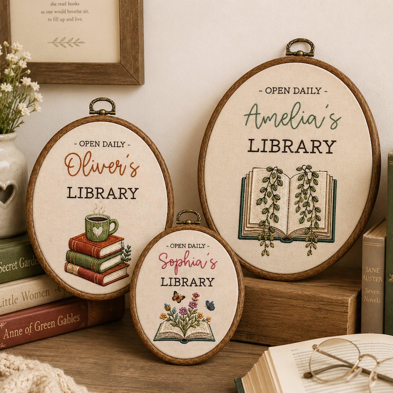 May include: Three oval embroidery hoops featuring personalized library designs on cream fabric. The hoops display the text 'OPEN DAILY' and names 'Oliver's', 'Amelia's', and 'Sophia's' above 'LIBRARY', accompanied by embroidered books, plants, and flowers.