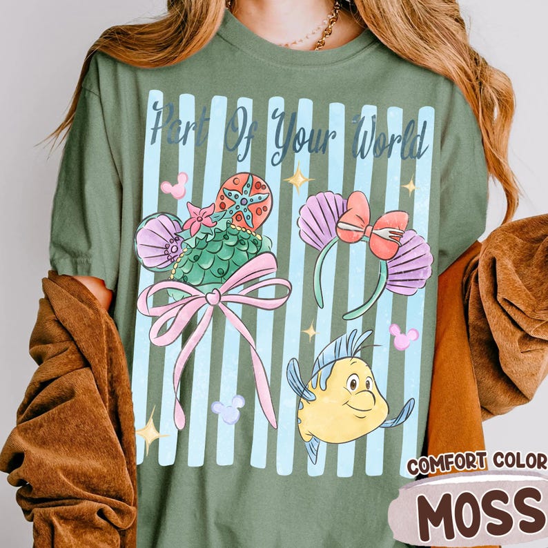 Puede incluir: Una camiseta verde salvia con el texto Part Of Your World sobre rayas verticales azul claro. El gr&aacute;fico incluye dos pares de orejas de rat&oacute;n, una con una estrella de mar y un lazo, la otra con conchas, y un pez amarillo con aletas azules.