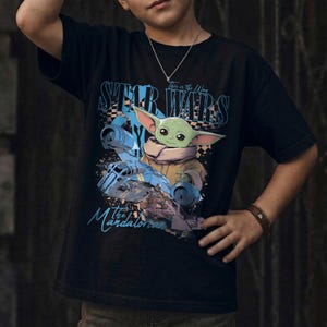 May include: Black t-shirt featuring a graphic of Grogu from Star Wars, with the text "STAR WARS" and "The Mandalorian." The design includes a blue spaceship and the phrase "This is the Way."