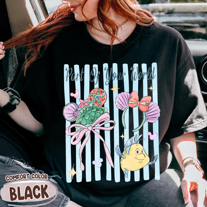 Puede incluir: Una camiseta negra con un estampado gr&aacute;fico de rayas verticales azul claro y el texto Part Of Your World. El dise&ntilde;o incluye una concha verde, una estrella de mar, un lazo rosa, una diadema con orejas y un personaje de pez amarillo.
