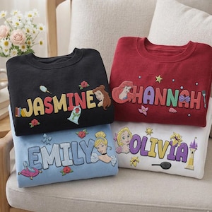 Puede incluir: Cuatro sudaderas en negro, rojo, azul claro y blanco, cada una bordada con un nombre y un gr&aacute;fico de princesa. La sudadera negra dice "JASMINE" con una princesa e iconos relacionados. La sudadera roja dice "HANNAH" con una princesa. La sudadera azul claro dice "EMILY" con una princesa. La sudadera blanca dice "OLIVIA" con una princesa.